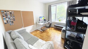 A room with a grey sofa, a desk with a computer, a window with a view of trees, a bulletin board, and a black shelving unit with a GVM logo.