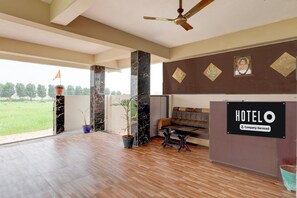 A hotel reception area with a sign indicating it's a company-serviced hotel, a ceiling fan, a sofa, and a view of a green field through large windows.