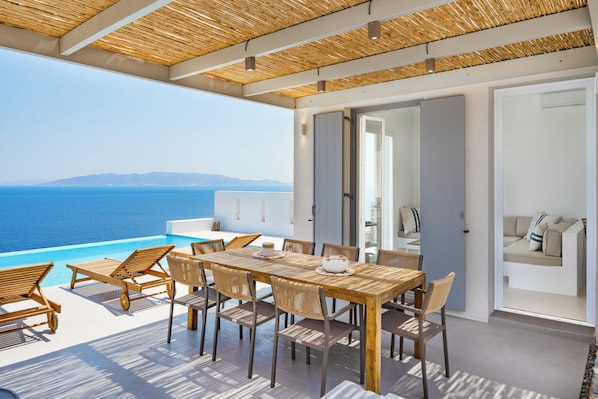 Eine sonnendurchflutete Terrasse mit Holztisch und -stühlen, Blick auf das Meer und ein modernes Interieur mit Sofa und einer Tür.