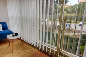 A balcony with white curtains, a blue sofa, a small table with a bowl, and a view of a parking lot and greenery. - Beautiful apartment with pool close to the beaches (Sainte-Maxime)