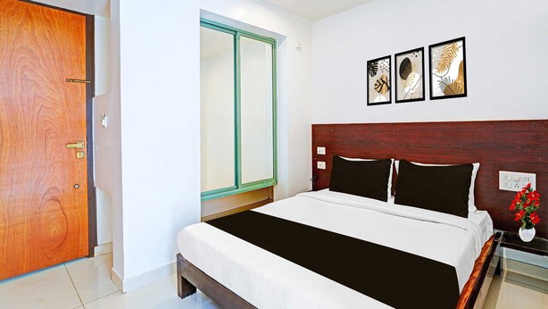 A hotel room with a wooden door, a bed with black and white bedding, a nightstand with a vase of red flowers, and three framed wall art pieces. - Classic With Twin Bed Kaveri Road Mysore (Mysore)