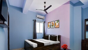 A bedroom with a bed, a ceiling fan, an air conditioning unit, a window with curtains, a table with a vase, and a mirror.