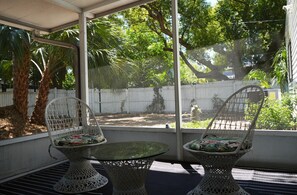 Eine überdachte Terrasse mit zwei Korbstühlen und einem Glastisch, mit Blick auf einen Garten mit weißem Zaun und Bäumen.