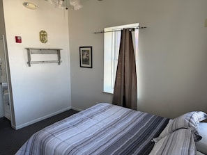 Ein Schlafzimmer mit Bett, einem Fenster mit Vorhängen, einem Wandregal und einem Bild.
