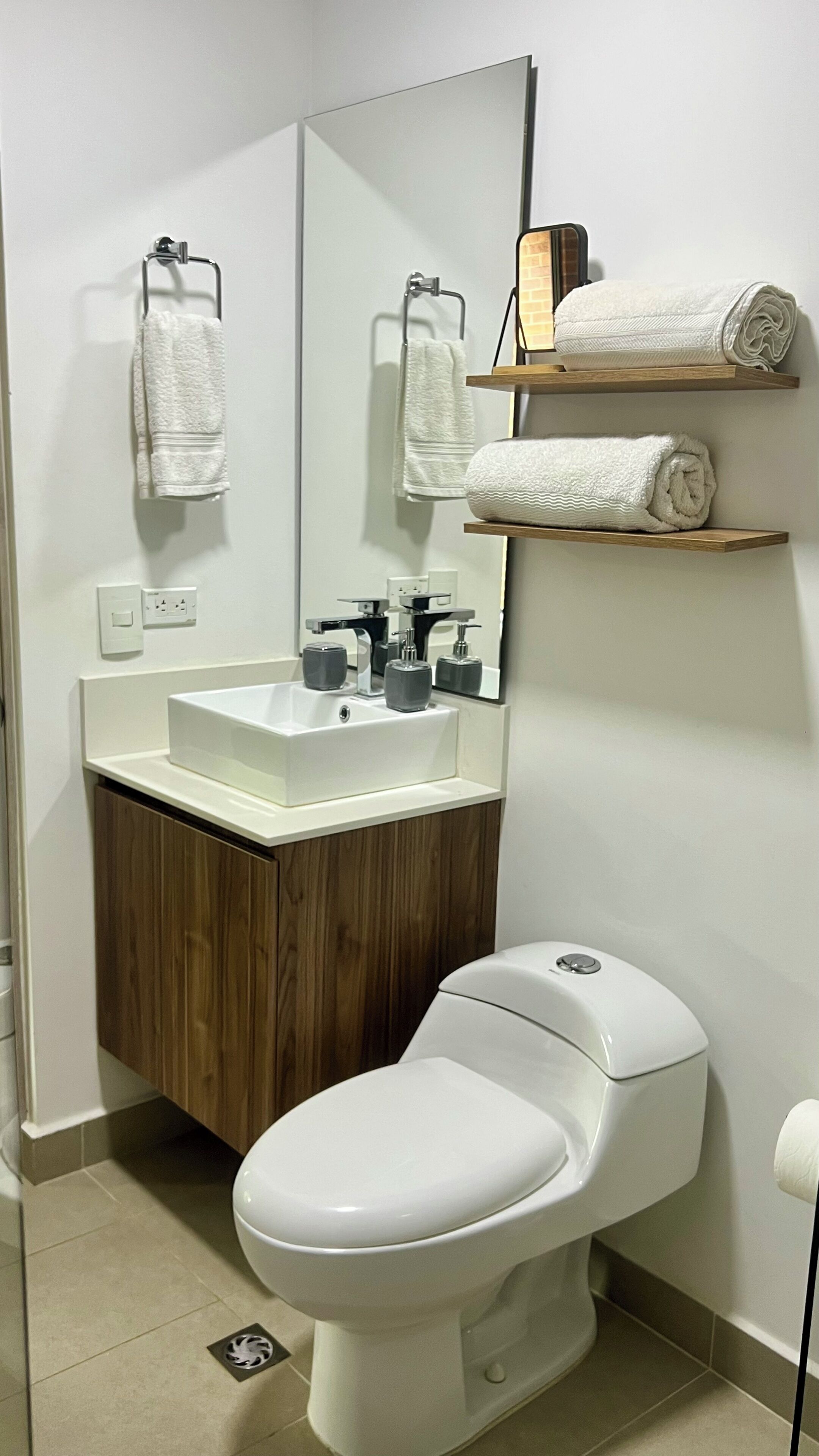 Une salle de bain moderne avec un lavabo, un miroir et des toilettes, le tout de couleur blanche. Des étagères en bois sont fixées au-dessus du lavabo et servent à ranger des serviettes pliées.