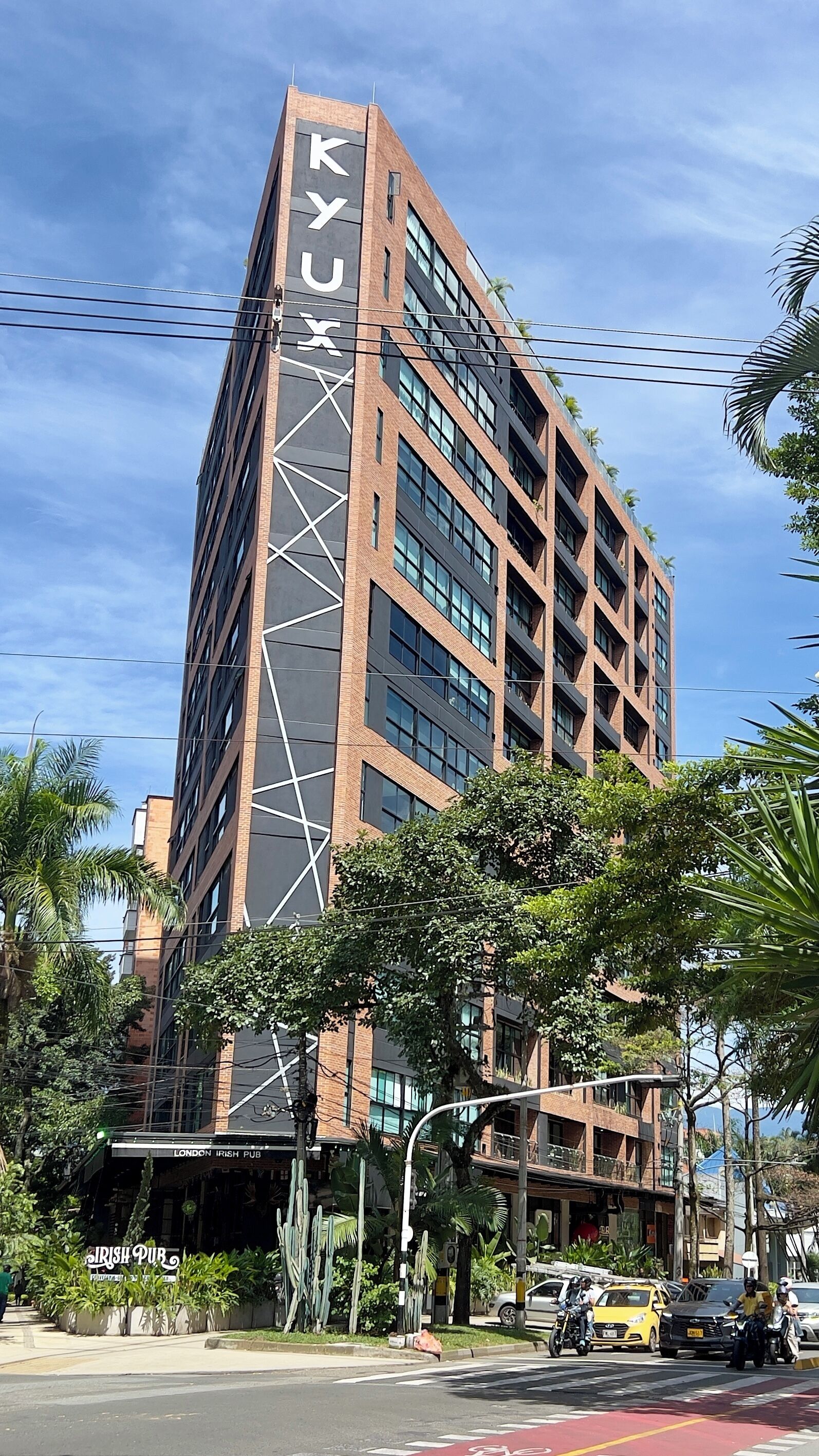 Un edificio alto y moderno con un llamativo letrero 'KYX', rodeado de vegetación y una calle con vehículos.
