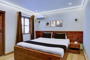 A hotel room with a wooden bed, a nightstand with a vase, an air conditioner, and framed artwork on the wall. - Classic With Balcony Traveller Guest (Calangute)