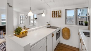 Une cuisine moderne avec des meubles blancs, un grand îlot central, des luminaires suspendus, une fenêtre offrant une belle vue et un panier en osier rempli de fruits.