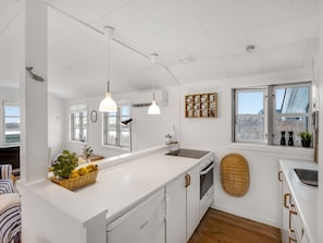 Una cocina moderna con armarios blancos, una isla grande, luces colgantes, un ventanal con vistas y una cesta de mimbre con fruta.