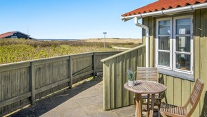 Une terrasse en bois avec une table et des chaises, une fenêtre surmontée d’un toit rouge, et une vue sur la plage et les dunes.