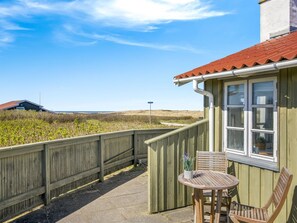 Una terraza de madera con una mesa y sillas, un ventanal con techo rojo y vistas a la playa y las dunas.