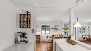 Une cuisine moderne équipée d’une machine à café, de rayonnages en bois où sont posées des tasses, d’un coin repas avec une table et des chaises, et offrant une vue sur l’extérieur grâce à une fenêtre.