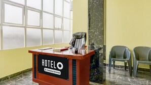 A hotel reception desk with a sign reading 'HOTEL' and 'Company-Serviced.' There are two plastic chairs to the right and a large window behind the desk.