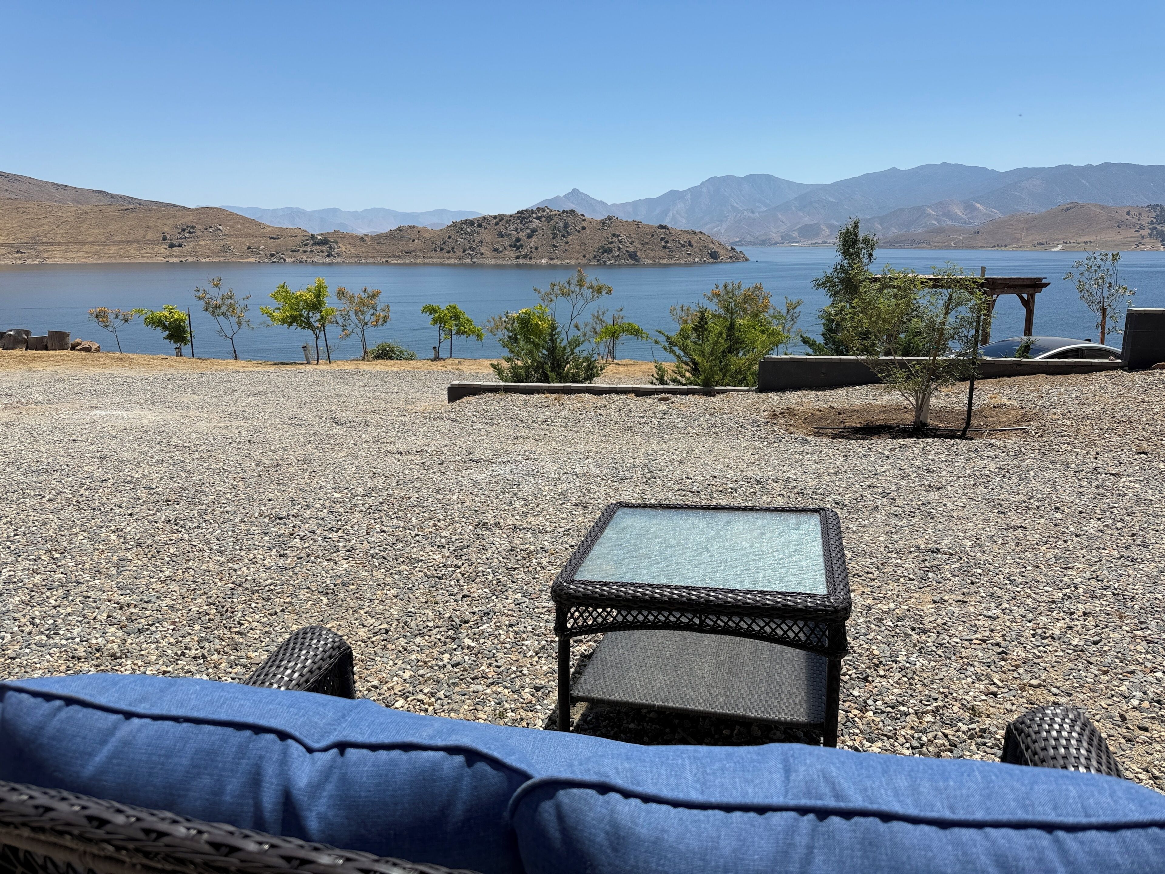 A gravel patio with a wicker chair and a small table, overlooking a lake with mountains in the background.