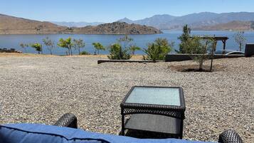 A gravel patio with a wicker chair and a small table, overlooking a lake with mountains in the background.