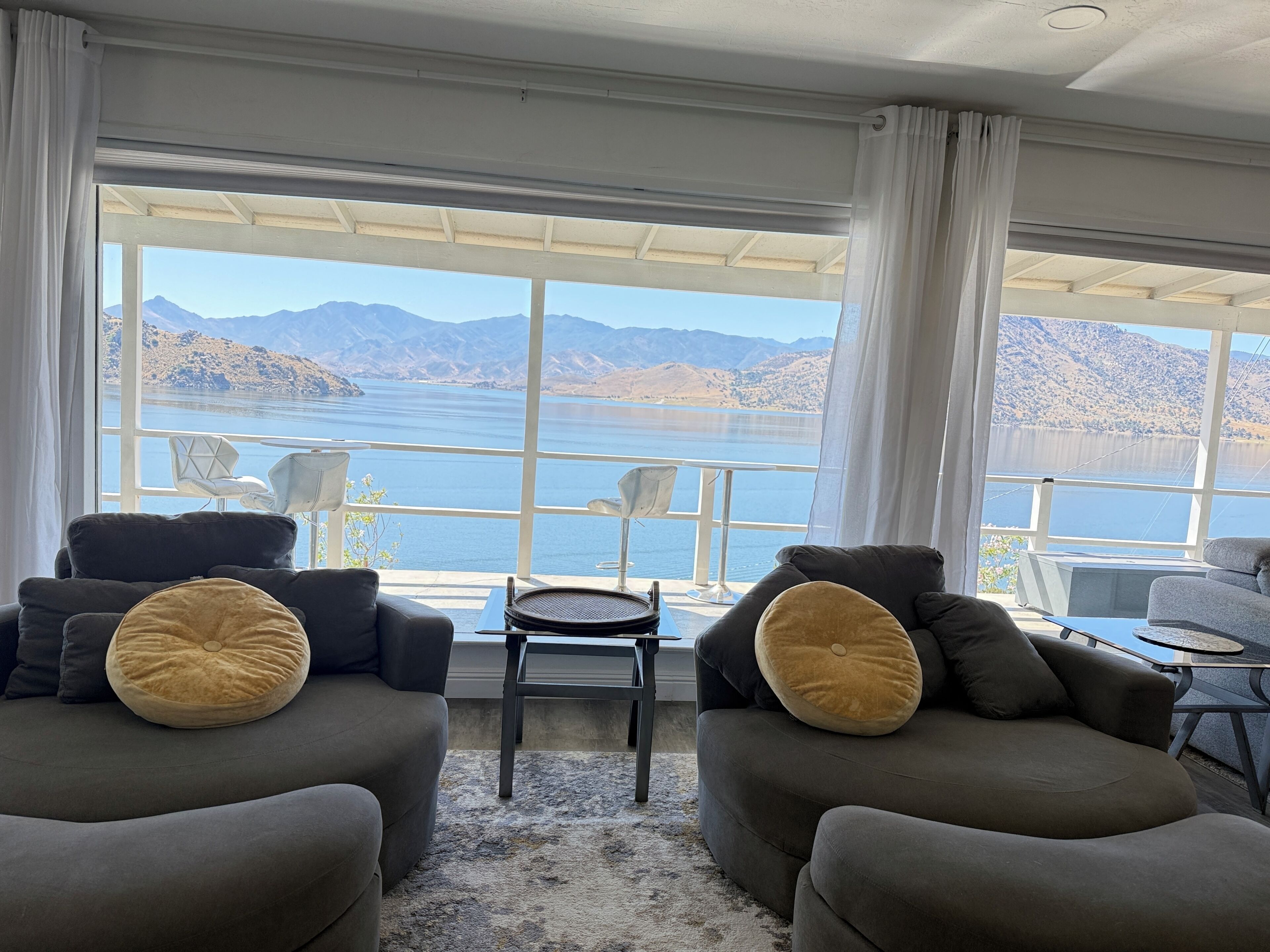Una sala moderna con dos sofás, una mesita y una silla, todos orientados hacia un gran ventanal con vistas a un lago y montañas.