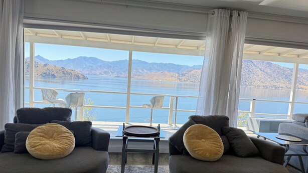 Una sala moderna con dos sofás, una mesita y una silla, todos orientados hacia un gran ventanal con vistas a un lago y montañas.