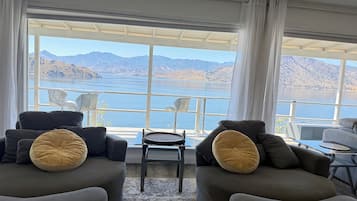Una sala moderna con dos sofás, una mesita y una silla, todos orientados hacia un gran ventanal con vistas a un lago y montañas.