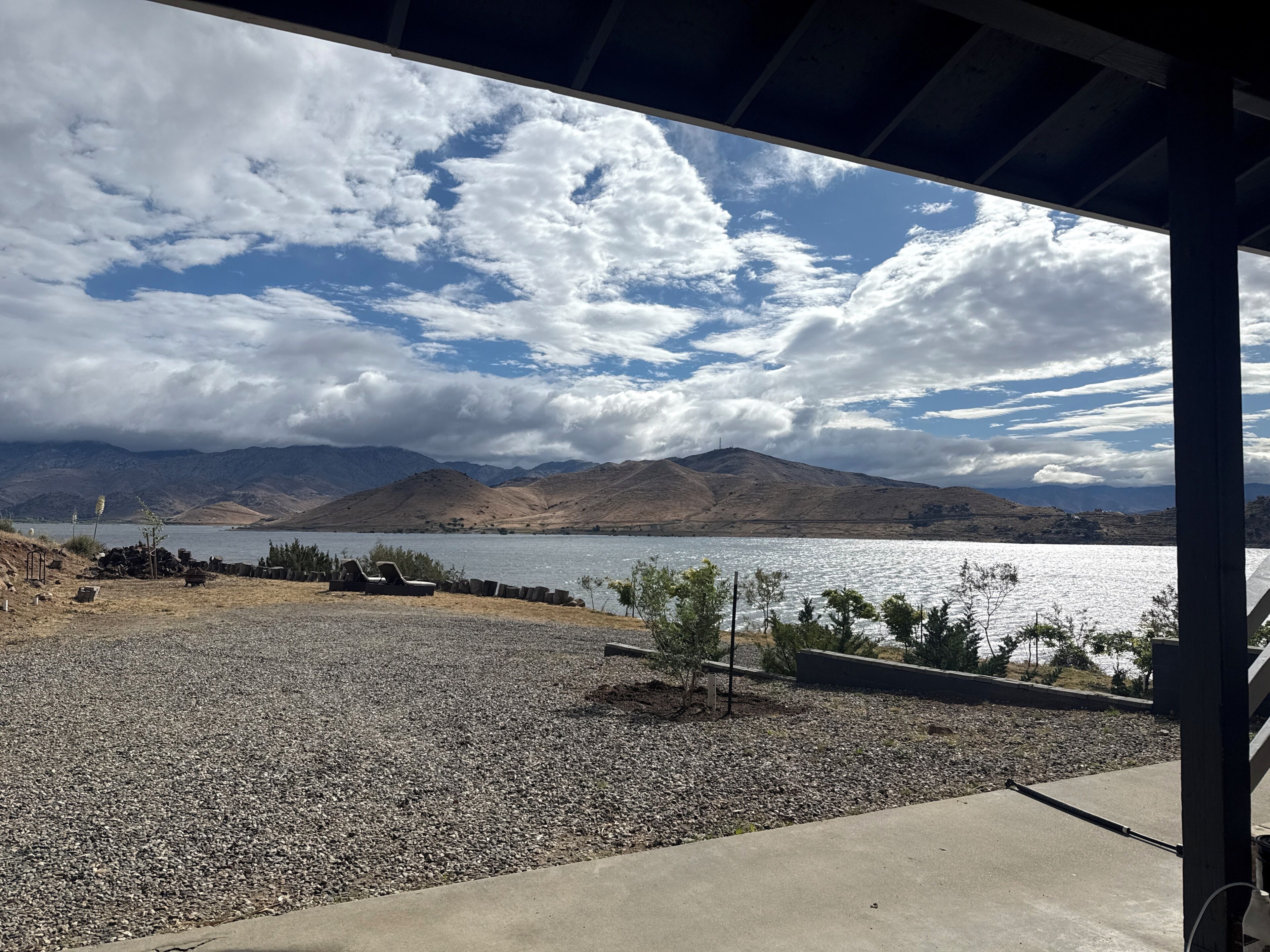 Vista de un lago con montañas al fondo, una zona de grava con una tumbona y una estructura cubierta.
