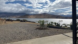 Vista de un lago con montañas al fondo, una zona de grava con una tumbona y una estructura cubierta.