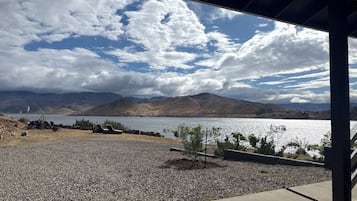 Vista de un lago con montañas al fondo, una zona de grava con una tumbona y una estructura cubierta.