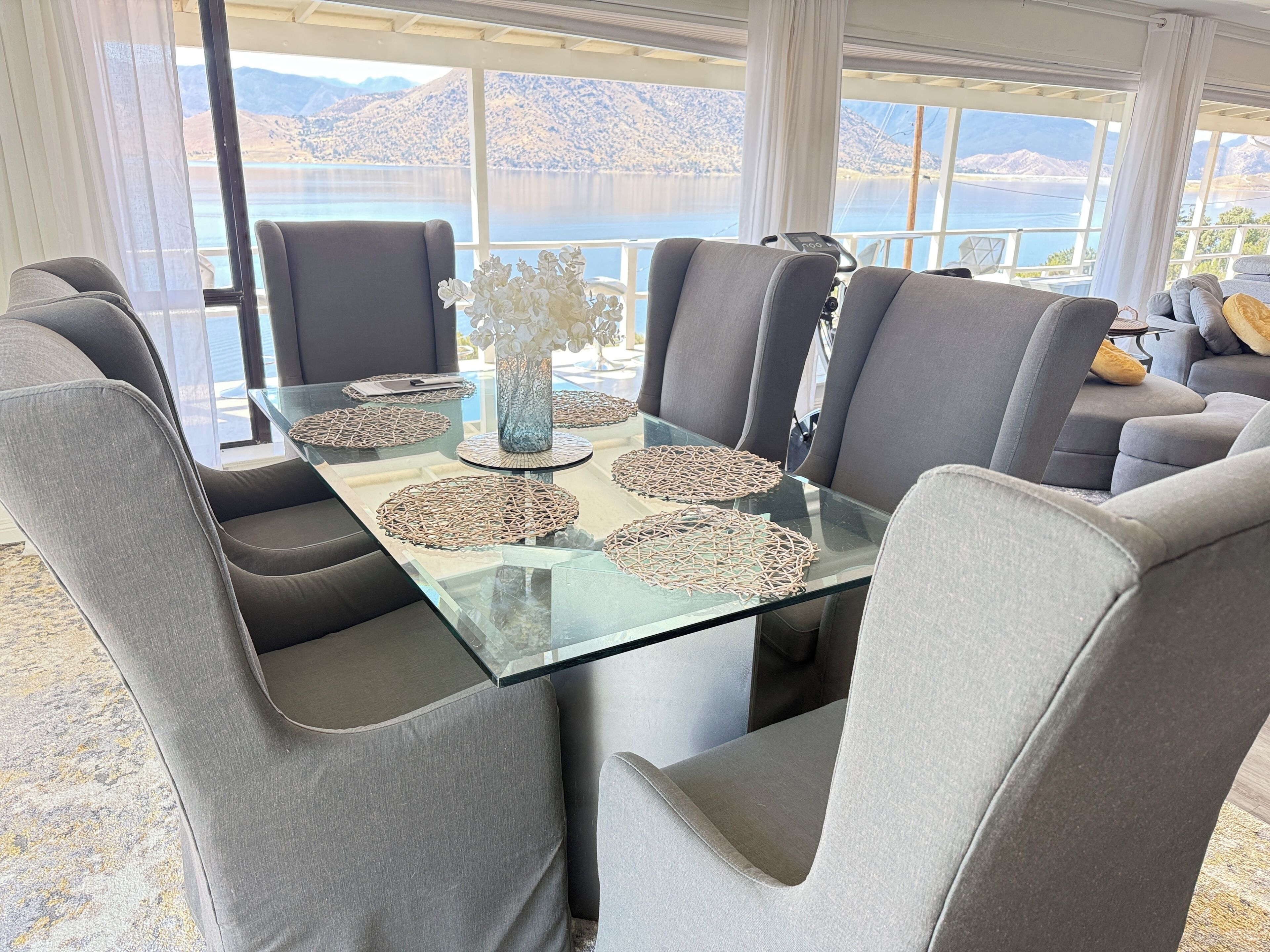 Uma área de jantar moderna com mesa de vidro, cadeiras estofadas cinzentas e um vaso com flores brancas.