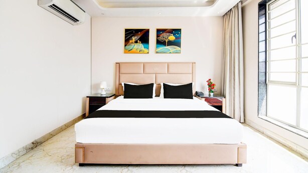 A hotel room with a large bed, a nightstand, a wall-mounted air conditioner, two paintings, and a large window with curtains. - Premium New Krish Residency (Barasat)