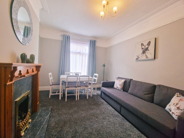 A living room with a fireplace, a dining table with chairs, a sofa, a chandelier, and a framed picture on the wall.