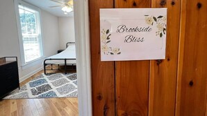 A bedroom with a bed, a nightstand, a dresser, a window with blinds, and a wooden door with a sign. - Brookside Bliss at The Washington Inn (Fries)