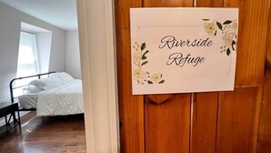 Een slaapkamer met een bed, een bankje en een raam. Op de deur staat een bordje met de tekst 'Riverside Refuge'.