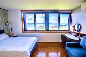 A hotel room with a bed, a small table, a blue armchair, and a large window with a view of the sea.