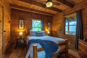 Une chambre dans une cabane en bois, avec un lit, des tables de chevet, un ventilateur de plafond et des fenêtres avec des rideaux.