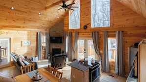 Une cabane avec une chambre à coucher située en mezzanine, un salon avec une cheminée, une cuisine équipée d’une cuisinière et de grandes fenêtres.