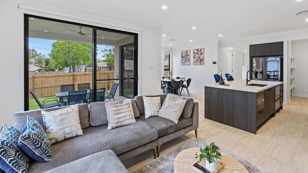 Un salon moderne avec un canapé gris, un coin repas comprenant une table et des chaises, une cuisine dotée d’un plan de travail foncé et une porte-fenêtre coulissante donnant sur un jardin.