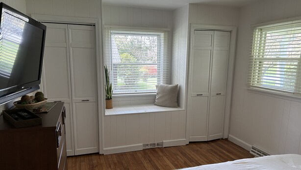 Quarto com cama, televisão, um assento junto à janela com uma planta e uma porta branca.