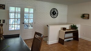 Um quarto com parede de tijolos brancos, um relógio preto redondo, uma mesa de jantar de madeira e um banco.