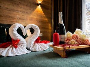 A wooden wall with a light fixture, a bottle of Crico wine, two swan-shaped towels, roses, and a wooden tray with red beads.