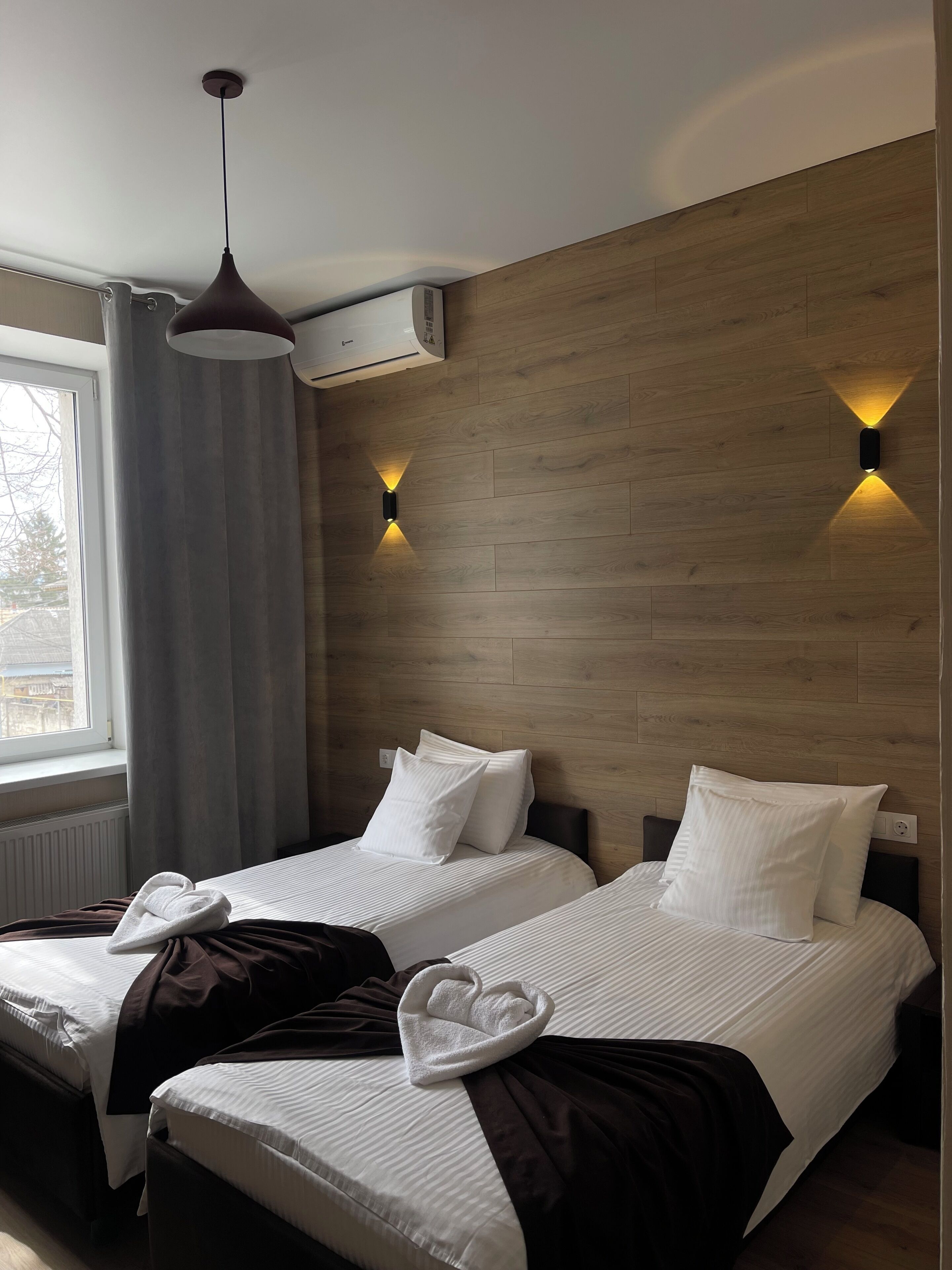 A hotel room with two beds, a window with curtains, an air conditioner, and wall-mounted lights.