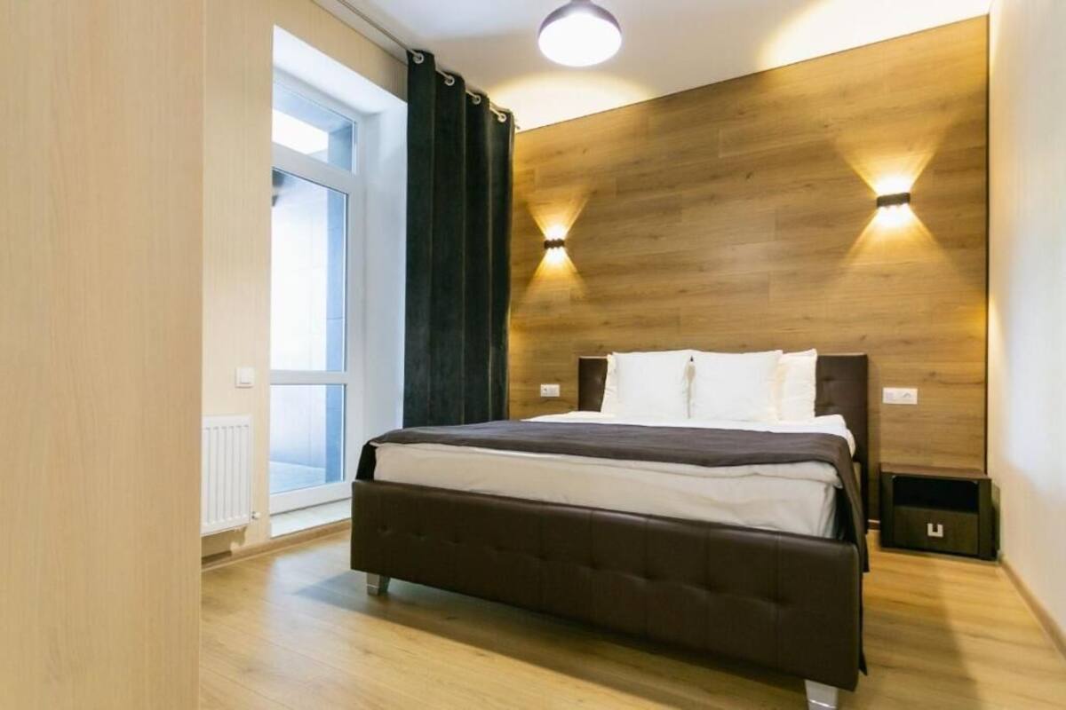 A bedroom with a large bed, wooden headboard, two wall-mounted lights, a window with curtains, and a radiator.