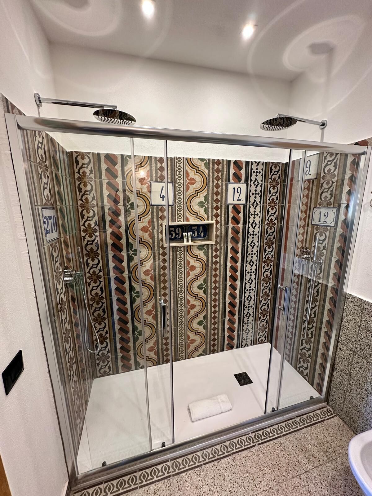 Un box de ducha con puerta de cristal, azulejos numerados y una toalla en el suelo.