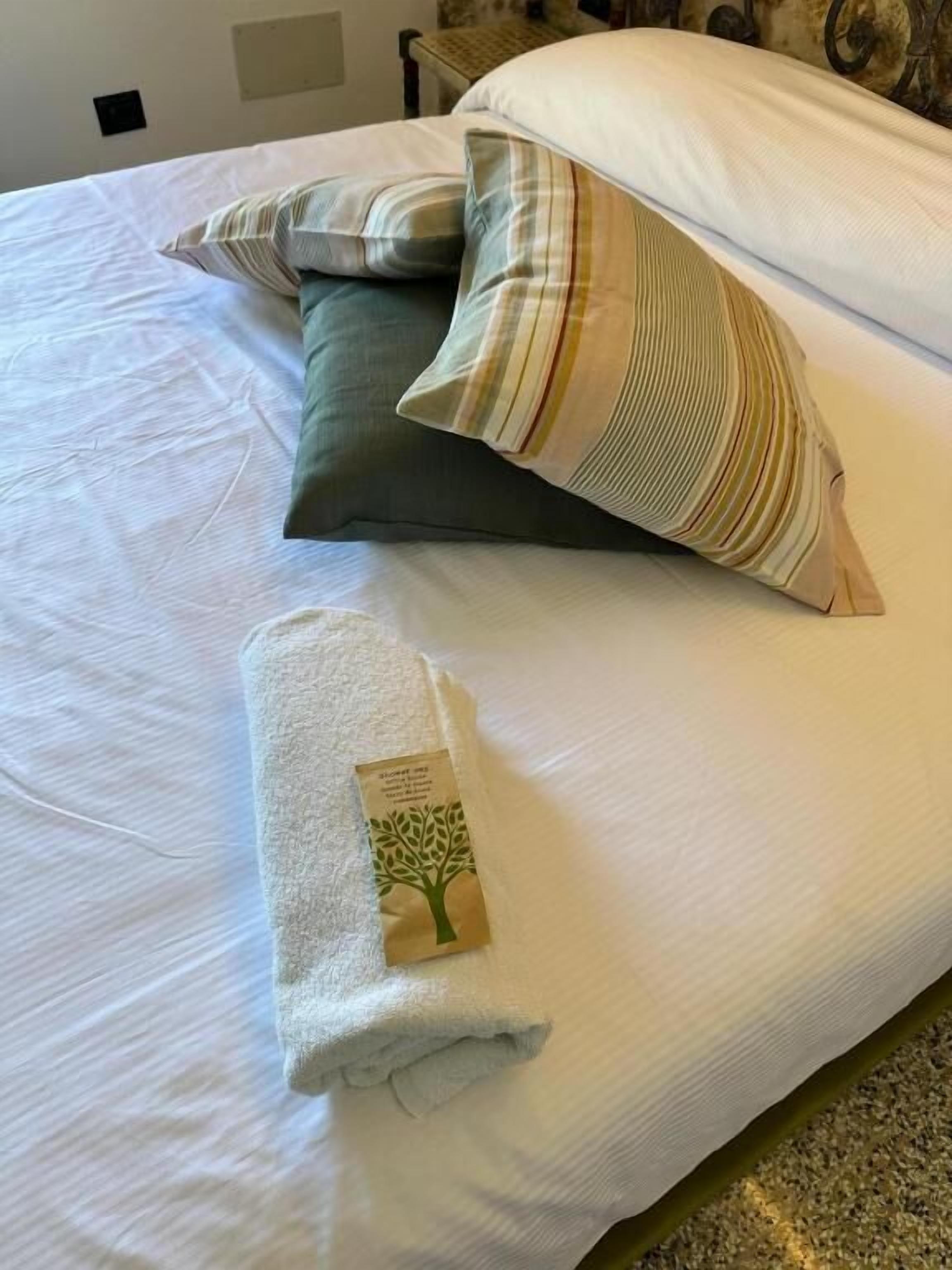 Ein ordentlich gemachtes Bett mit weißer Bettdecke, ein gefaltetes Handtuch mit Baum-Motiv und gestreifte Kissen.