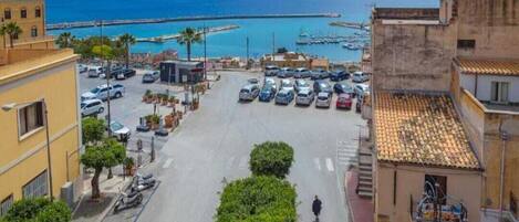 A coastal street with parked cars, a pedestrian, and buildings with balconies.