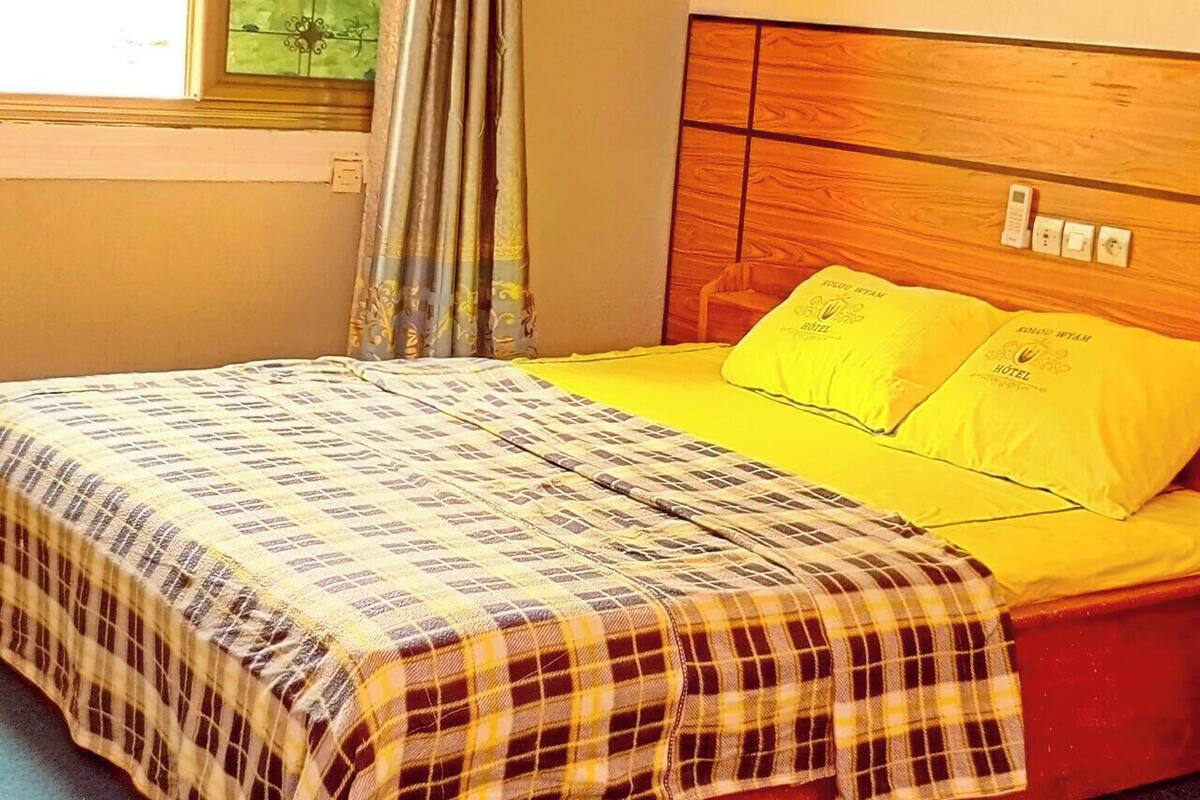 Un lit avec une couvre-lit à carreaux, un oreiller jaune et une tête de lit ornée d’un logo. Il y a une fenêtre avec des rideaux, un appareil de climatisation placé au-dessus, et un tableau accroché au mur.