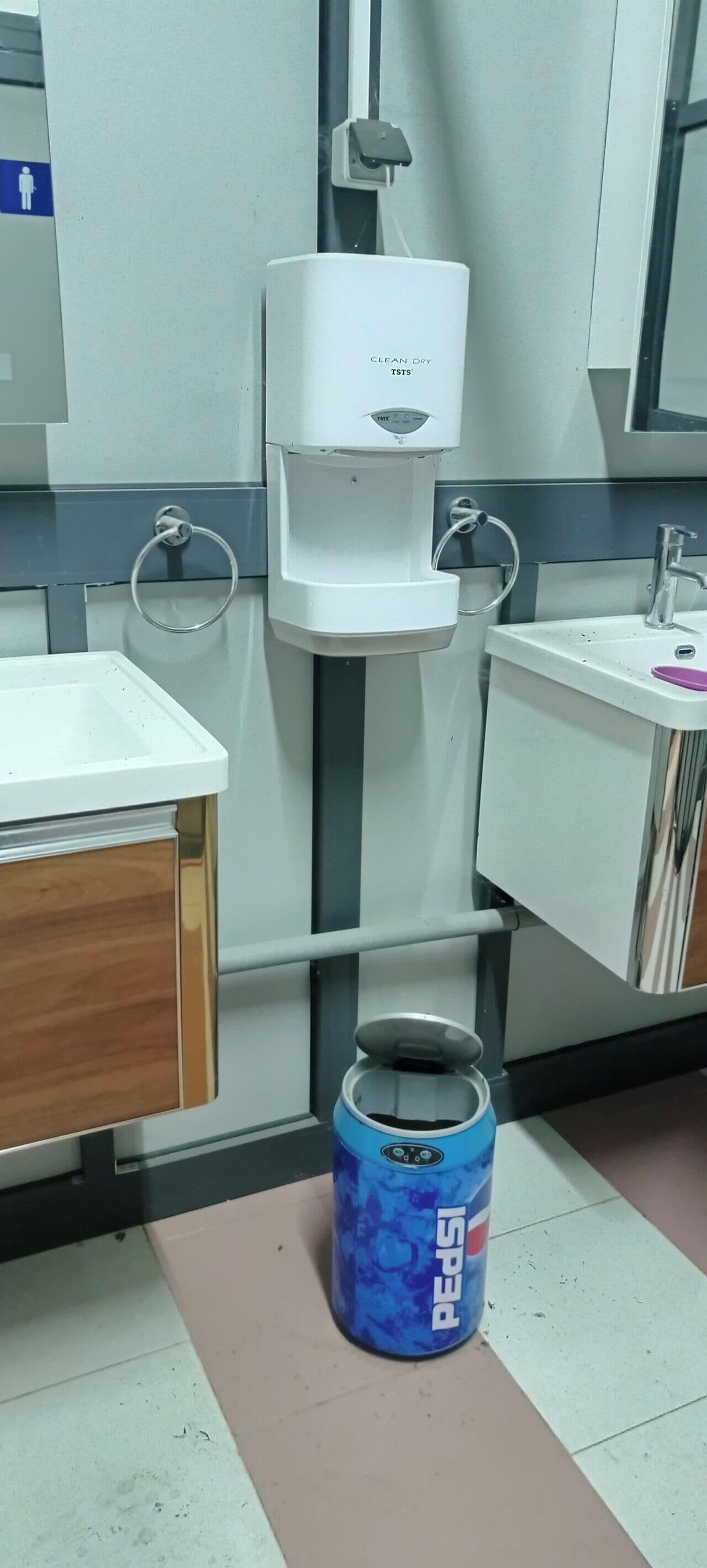 A restroom with a white sink, a mirror, a dispenser, and a blue Pepsi can on the floor.