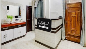 A hotel reception area with a marble-tiled floor, a reception desk, a wooden door, a clock, and a mirror with a plant. - Deluxegopal Singh Niwas (Varanasi)