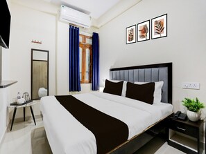 A hotel room with a bed, a bedside table with a phone and a plant, a mirror, a window with curtains, and an air conditioning unit. - Deluxegopal Singh Niwas (Varanasi)