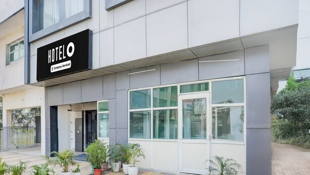 A hotel exterior with a sign reading 'HOTELO Company-Serviced,' large windows, and potted plants in front. - Saver Double Zirakpur JB Plaza (Dera Bassi)