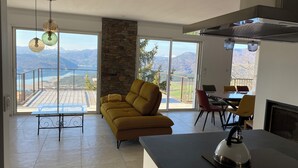 Una sala de estar moderna con sofá, mesa de comedor y área de cocina, con amplias ventanas que ofrecen vistas a la montaña.