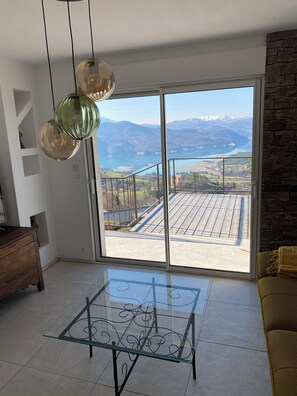 Una habitación con una mesa de vidrio, un aparador de madera, un sofá, una pared de piedra y vistas a montañas y un lago a través de grandes puertas de vidrio.