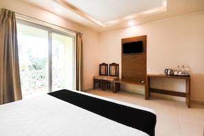 A hotel room with a bed, a TV mounted on the wall, a small table with a kettle, and a balcony with curtains.
